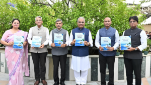 Woman in pink saree and five men holding National Coastal Mission 2.0 reports standing before green foliage government