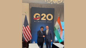 US flag left, Piyush Goyal shaking hands with a man before a blue G20 Milwaukee 2026 banner, Indian flag right meeting.